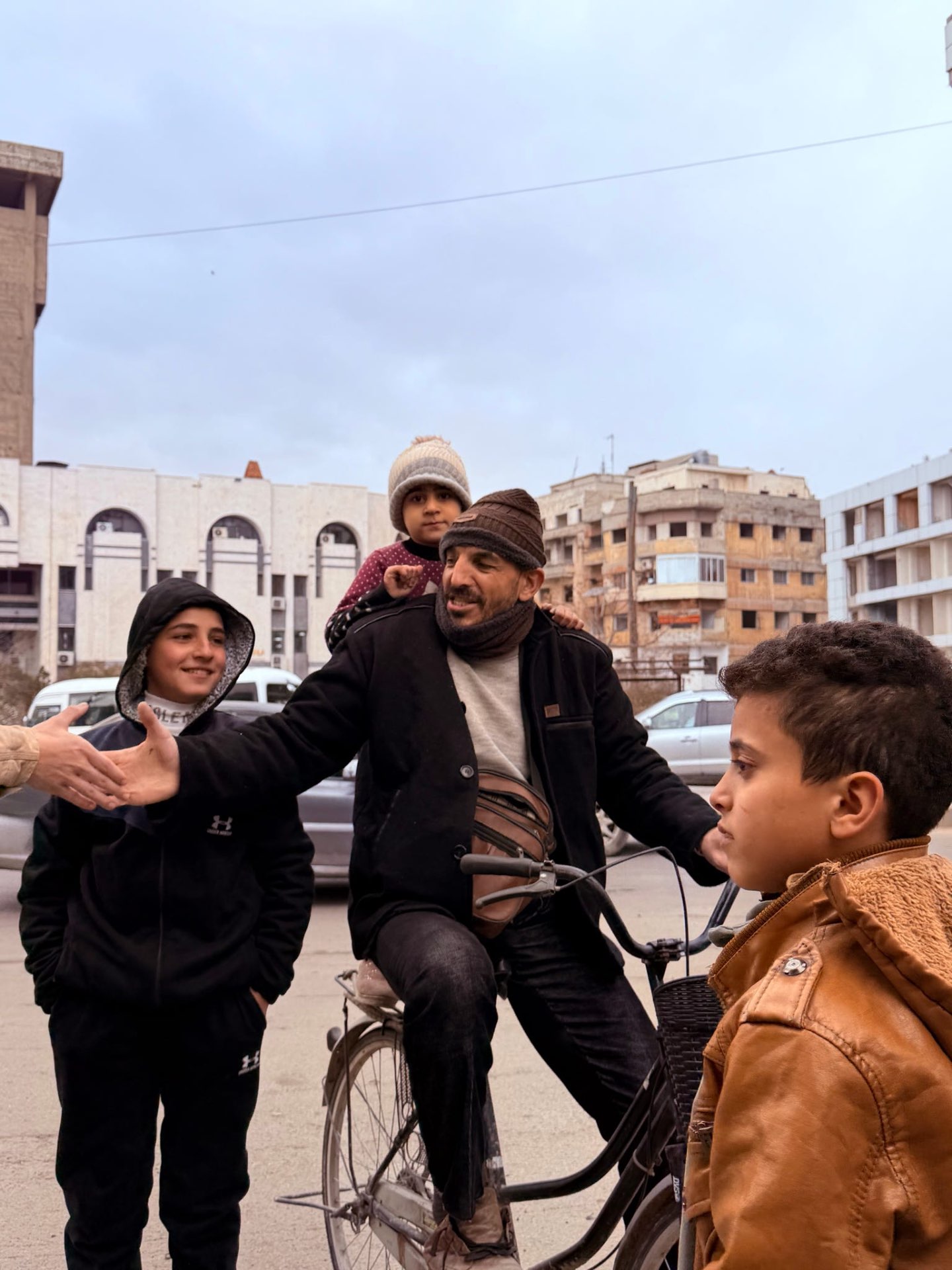 A man on a bicycle with a small child on his shoulders, greeting neighborhood kids in a Syrian street