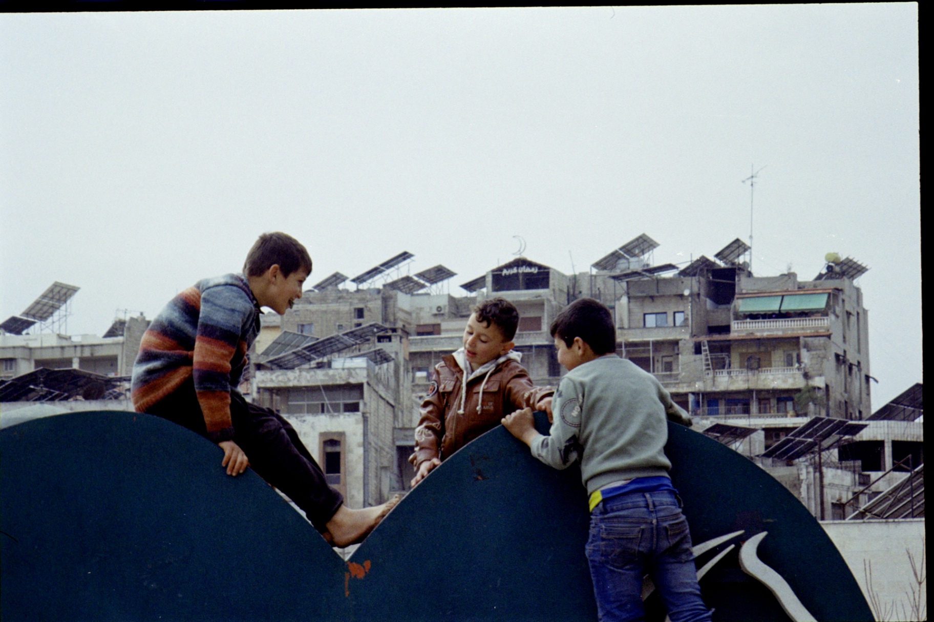 Three children playing on a rooftop sculpture above a Syrian neighborhood